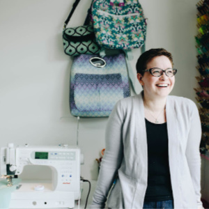 Samantha Hussey aka Mrs H from Sewing Patterns by Mrs H, in a sewing studio. Samantha is a light-skinned woman with short brown hair, wearing glasses as well as a grey cardigan over a black top and blue jeans. Some of her bag designs hang on the wall behind her.